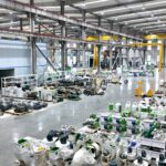 Wide pellet equipment workshop showing production areas, machines, parts, cranes, and staff